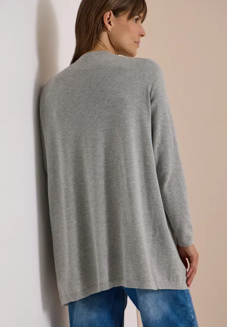 Offener Cardigan in langer Passform mineral grey melange Frau von hinten in grauem, locker fließendem Strickcardigan mit blauen Jeans, lehnt an hellem Hintergrund.
