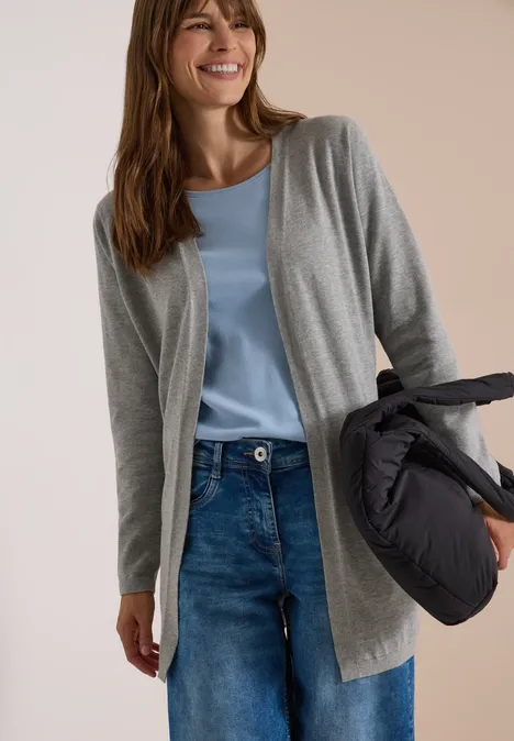Offener Cardigan in langer Passform mineral grey melange Eine lächelnde Frau trägt graue Strickjacke, hellblaues Top, Jeans und hält eine Daunenjacke.