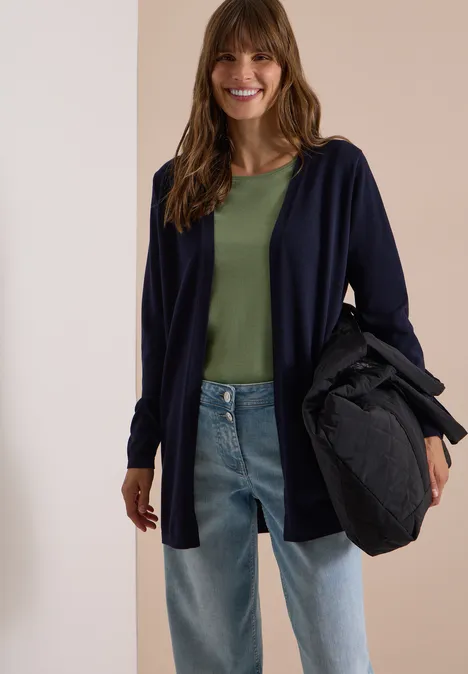 Een lachende vrouw draagt een groen shirt, een donkerblauwe cardigan, spijkerbroek en houdt een zwarte tas vast.