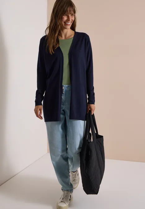 Vrouw met lang marineblauw vest, groen topje, losse jeans en sneakers, draagt een grote zwarte tas.