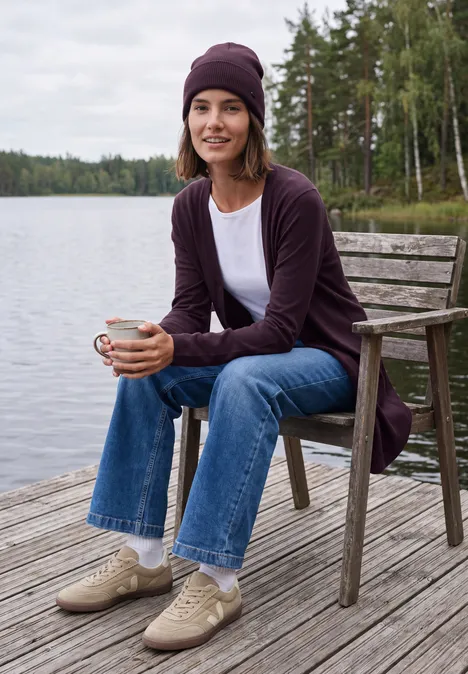 Femme en jean et cardigan au bord d’un lac, assise sur un quai en bois, tient une tasse et sourit.