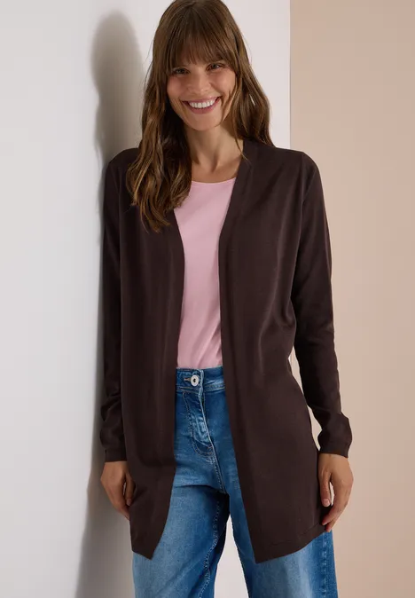 Vrouw lacht en leunt tegen muur, roze shirt, bruin vest en blauwe jeans.