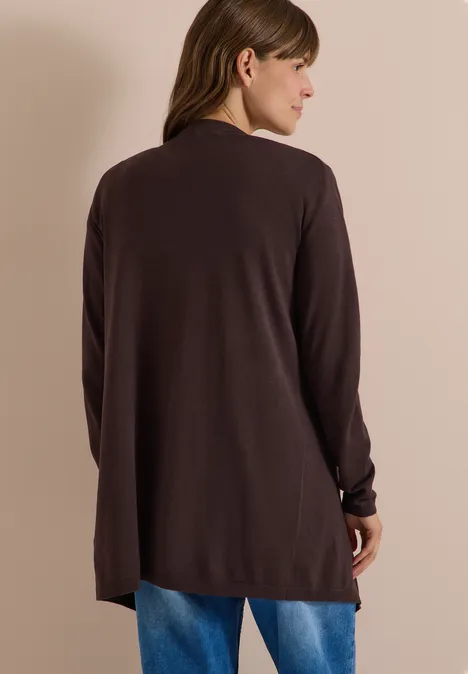 Vrouw van achteren in een lange donkere cardigan en blauwe spijkerbroek tegen neutrale achtergrond.