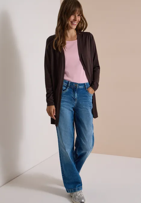 Lachende vrouw in blauwe jeans, roze top en zwart vest voor een neutrale achtergrond.