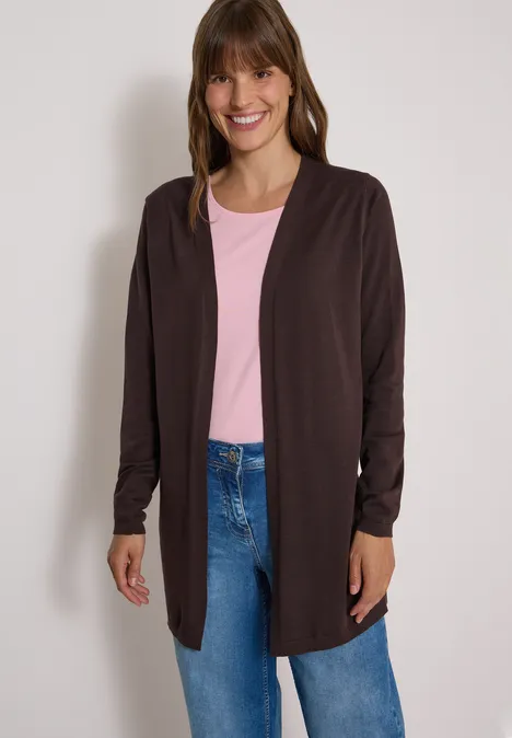 Een lachende vrouw draagt een donkerbruine open cardigan, een roze t-shirt en een blauwe jeans.