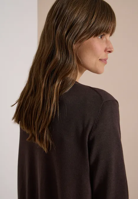 Zijaanzicht van een vrouw met golvend bruin haar in een donkerbruine top tegen een beige achtergrond.
