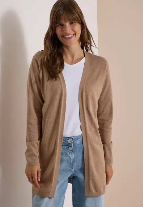 Een glimlachende vrouw draagt een beige open vest, wit T-shirt en jeans voor een lichte achtergrond.