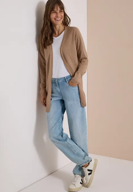 Een vrouw leunt tegen een muur met een wit shirt, lichtblauwe jeans, beige vest en witte sneakers.