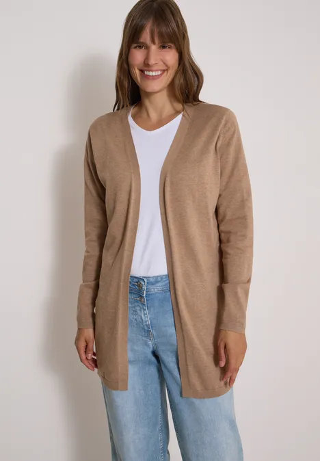 Vrouw in beige vest, wit shirt en spijkerbroek lacht voor een lichte muur.