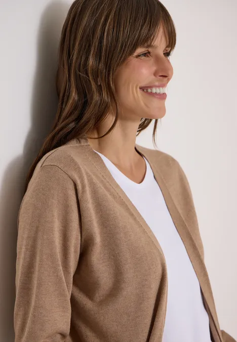 Vrouw in beige vest en wit T-shirt leunt glimlachend tegen een muur, zijaanzicht.
