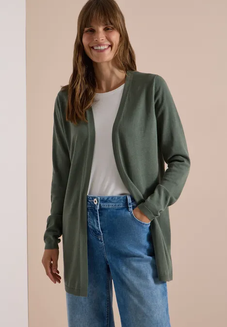 Eine lächelnde Frau in grünem Cardigan, weißem Shirt und blauen Jeans vor neutralem Hintergrund.