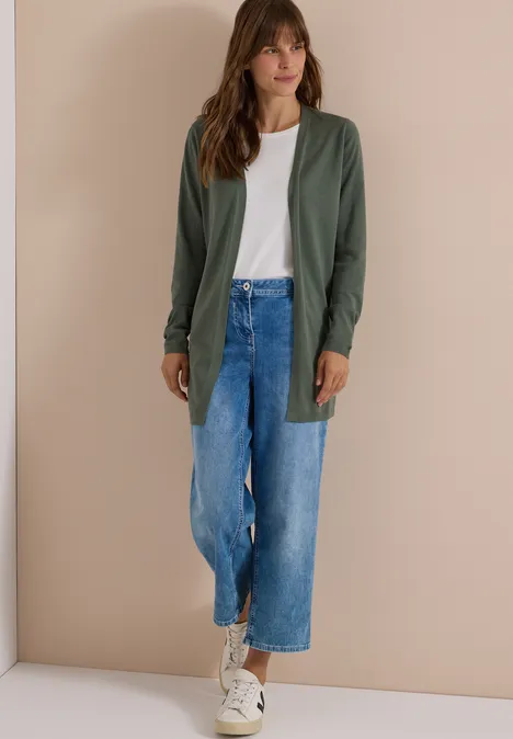 Frau in weißem T-Shirt, grüner Strickjacke, weitem blauen Jeans-Culotte und weißen Sneakern vor beiger Wand.