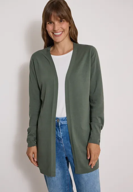 Frau in grünem Cardigan, weißem T-Shirt und Jeans, sie lächelt vor hellem Hintergrund.