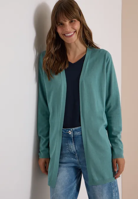 Junge Frau lehnt lächelnd an einer Wand, trägt grünen Cardigan, dunkelblauen Shirt und Jeans.