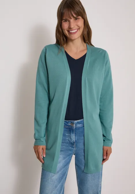 Frau im tealfarbigen Cardigan, dunkelblauem Shirt und Jeans, lächelt vor hellem Hintergrund.