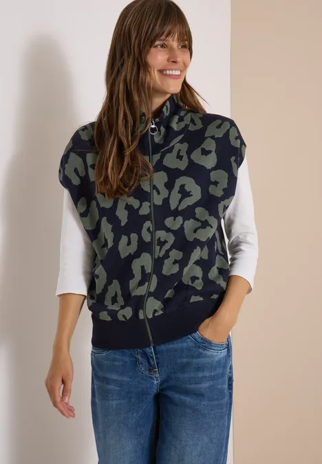 Frau in Jeans und weißem Shirt mit ärmelloser marine-grüner Weste im Leopardenmuster, sie lächelt.