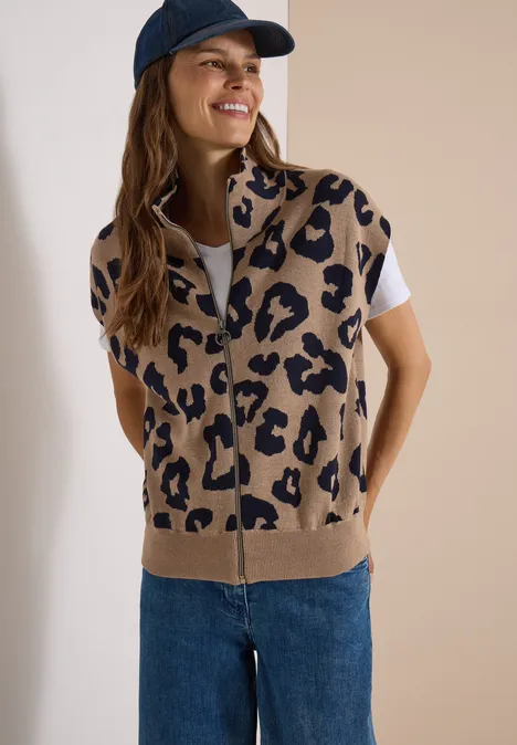 Frau mit beigem Strick-Gilet im Leopardenmuster, weißem Shirt, blauen Jeans und navy Cap lächelnd.