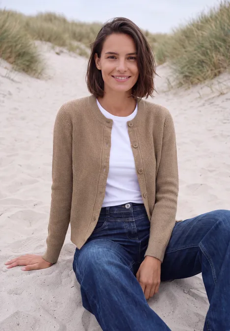 Vest met ronde hals en knoopsluiting creamy mocha melange Een glimlachende vrouw in spijkerbroek en vest zit ontspannen op duinen bij het strand.