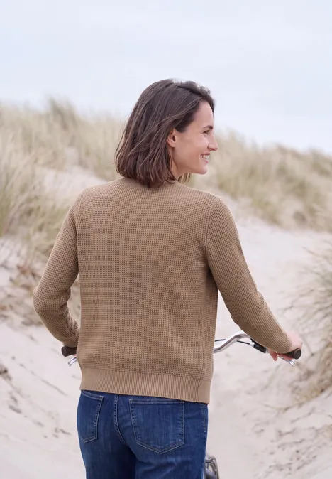 Vest met ronde hals en knoopsluiting creamy mocha melange Een vrouw in een bruine trui en spijkerbroek duwt een fiets over de duinen, omringd door helmgras.