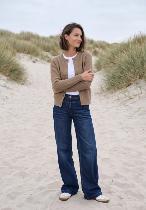 Vest met ronde hals en knoopsluiting creamy mocha melange Een vrouw in spijkerbroek en vest staat ontspannen tussen duingras op het strand.