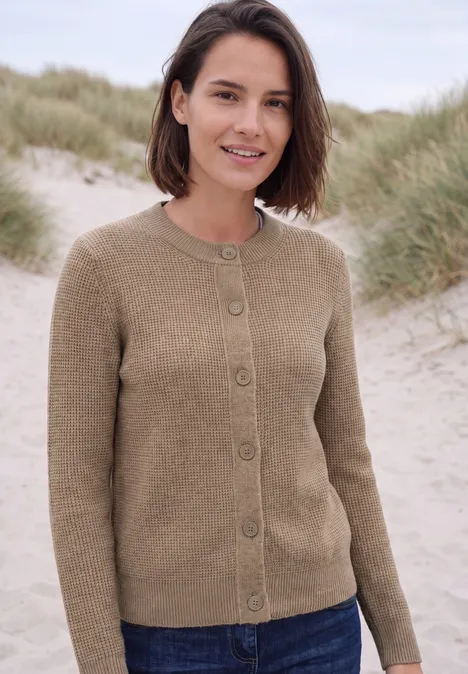Vest met ronde hals en knoopsluiting creamy mocha melange Vrouw met kort bruin haar in beige vest, ontspannen staand op een duinenstrand.