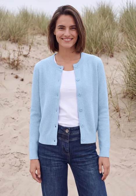 Vest met ronde hals en knoopsluiting cosmopolitan blue melange Een glimlachende vrouw in een lichtblauw vest, wit topje en spijkerbroek staat voor grasduinen.