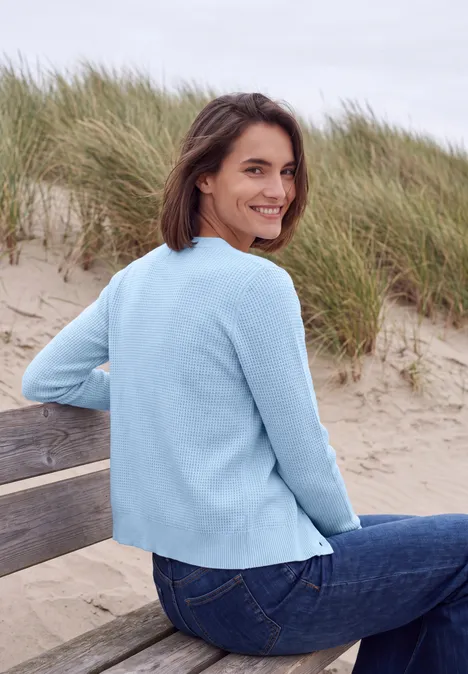 Vest met ronde hals en knoopsluiting cosmopolitan blue melange Een vrouw in een lichtblauwe trui zit glimlachend op een strandbank voor duinen.