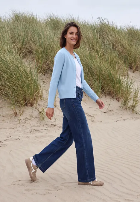 Vest met ronde hals en knoopsluiting cosmopolitan blue melange Een vrouw in jeans, wit shirt en lichtblauwe cardigan wandelt over de duinen op het strand.
