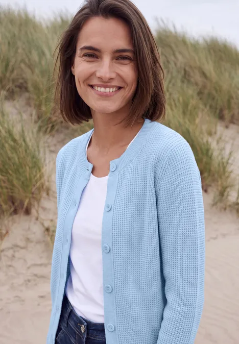Vest met ronde hals en knoopsluiting cosmopolitan blue melange Een glimlachende vrouw in een blauw vest voor duinen op het strand.