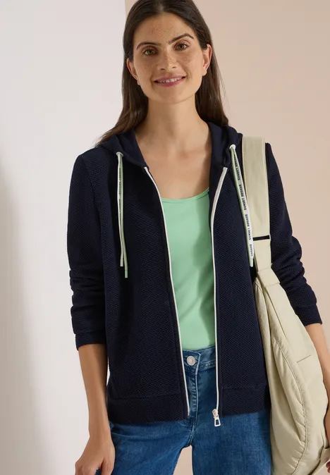 Lachende vrouw in donkerblau hoodie, mintgroen topje en spijkerbroek met een beige schoudertas.
