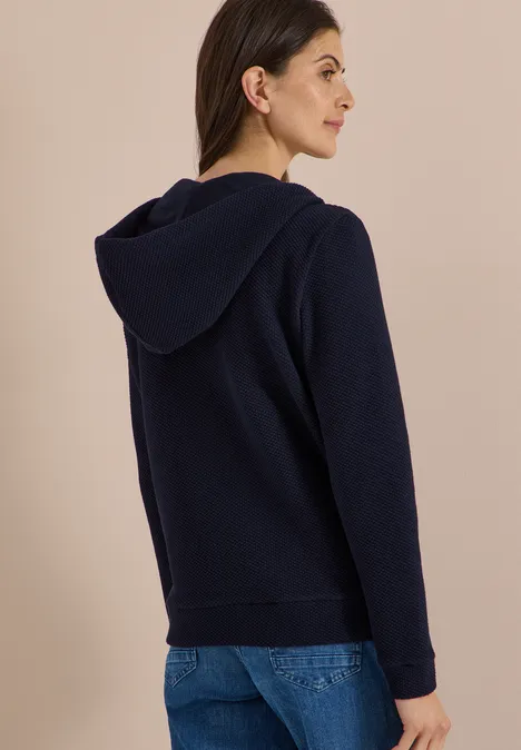 Vrouw van achteren in een donkerblauwe capuchontrui en spijkerbroek voor een beige muur.