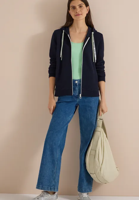 Jonge vrouw met marineblauwe hoodie, mintgroen topje, wijde jeans en grote linnen tas voor een beige muur.