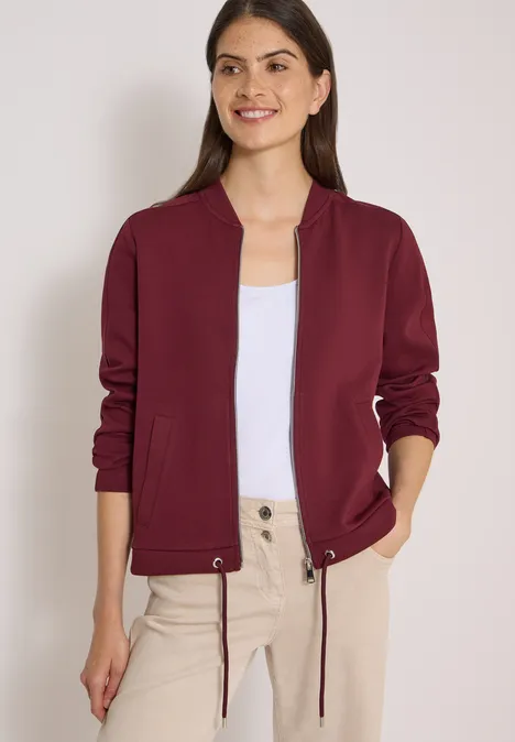 Sweat Blouson mit Tunnelzug red wine Frau trägt burgunderrote Bomberjacke über weißem Top und beigen Hosen.