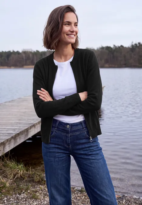 Langarm Cardigan mit Rundhals und Zipper Black Frau in Jeans, weißem Shirt und dunklem Cardigan steht lächelnd mit verschränkten Armen am Seeufer.
