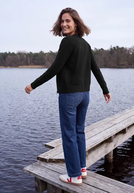 Langarm Cardigan mit Rundhals und Zipper Black Eine Frau steht auf einem Steg am See, dreht sich lächelnd zur Kamera, trägt schwarzen Pullover und Jeans.