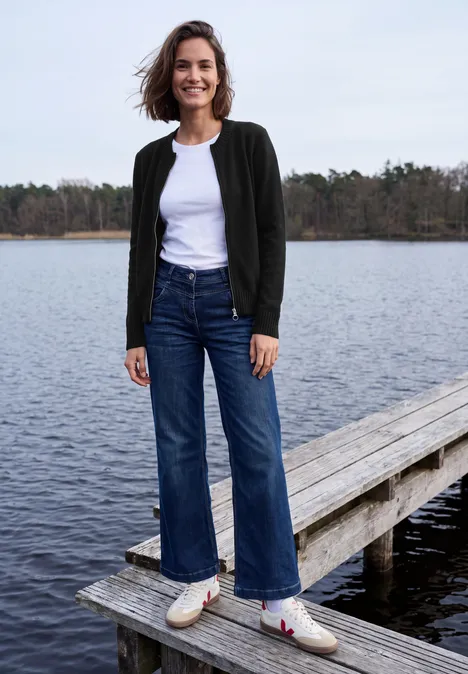 Langarm Cardigan mit Rundhals und Zipper Black Frau steht lächelnd auf Steg am See, trägt weißes Shirt, dunkle Jacke, weite Jeans und weiße Slip-ons.