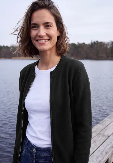 Langarm Cardigan mit Rundhals und Zipper Black Eine lächelnde Frau in weißem T-Shirt und dunkelgrünem Cardigan steht auf einem Steg am See.