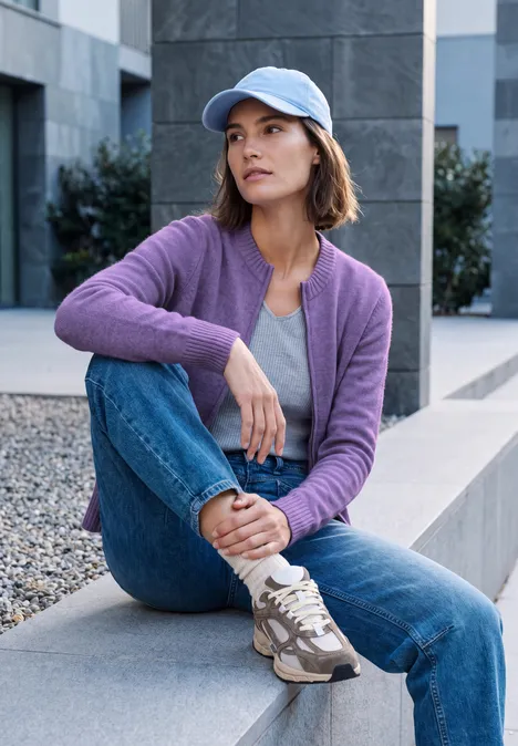 Cardigan zippé à manches longues et col rond pastel lilac melange Junge Frau sitzt in lila Strickjacke, Jeans und Baseballcap auf moderner Bank im urbanen Umfeld.