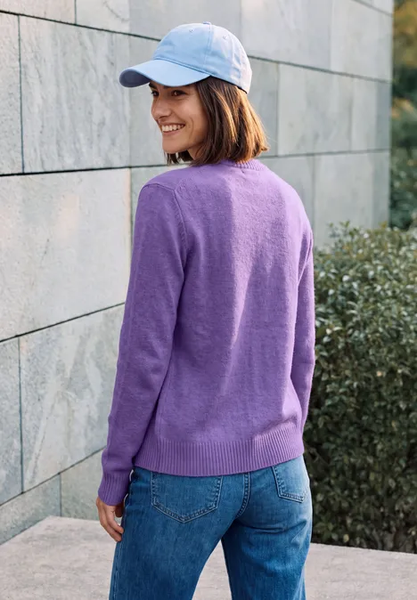 Cardigan zippé à manches longues et col rond pastel lilac melange Frau in lila Pullover, Jeans und weißer Mütze lächelt seitlich vor einer Steinwand.