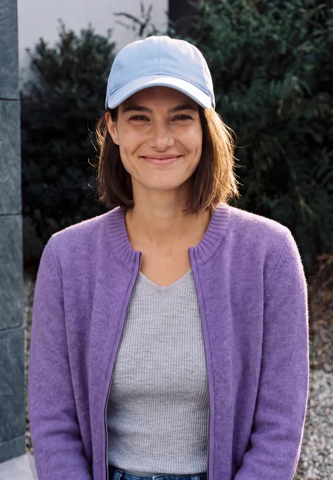 Cardigan zippé à manches longues et col rond pastel lilac melange Eine lächelnde Frau in fliederfarbener Strickjacke, grauem Shirt und hellblauer Baseballkappe steht draußen.