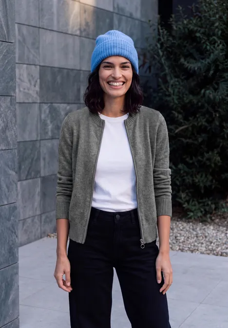 Langarm Cardigan mit Rundhals und Zipper flint grey melange Eine lächelnde Frau in blauer Mütze, weißem Shirt, grüner Strickjacke und schwarzer Hose vor einer Steinwand.