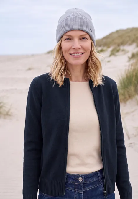 Frau mit grauer Mütze, schwarzer Strickjacke und hellem Pullover, die am Strand lächelnd in die Kamera schaut.