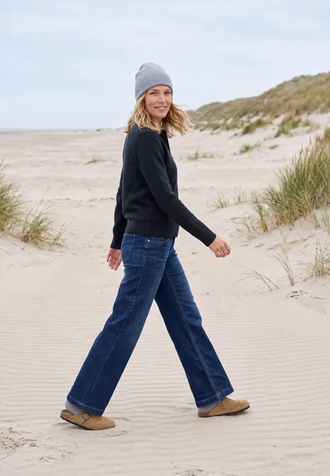 Eine Frau spaziert entspannt über Sanddünen am Meer, trägt Jeans, Pullover, Mütze und bequeme Schuhe.