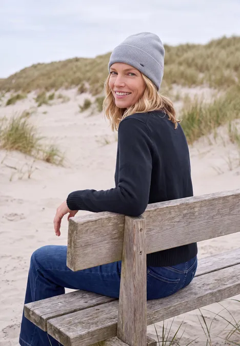 Eine Frau mit grauer Mütze und dunklem Pulli sitzt lächelnd auf einer Bank am Dünenstrand.