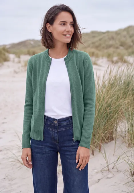 Vest met ronde hals en rits jewel green melange Een vrouw met groen jasje, wit t-shirt en jeans lacht op het strand voor duinen.
