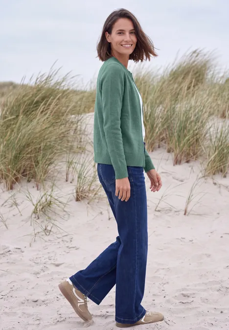Vest met ronde hals en rits jewel green melange Een glimlachende vrouw met groene trui en jeans staat op een strand voor duinen met helmgras.