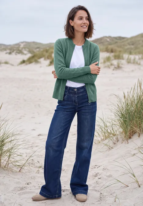 Vest met ronde hals en rits jewel green melange Een glimlachende vrouw staat op een zandstrand voor duinen, draagt een groen vest, wit shirt en wijde jeans.