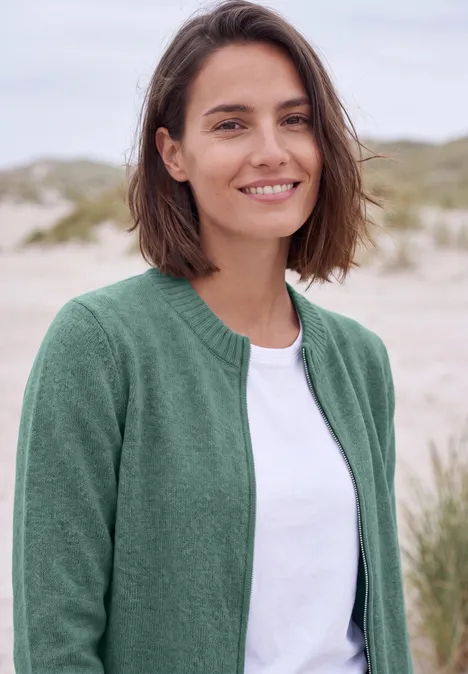 Vest met ronde hals en rits jewel green melange Een glimlachende jonge vrouw in een groen vest en wit shirt op het strand.