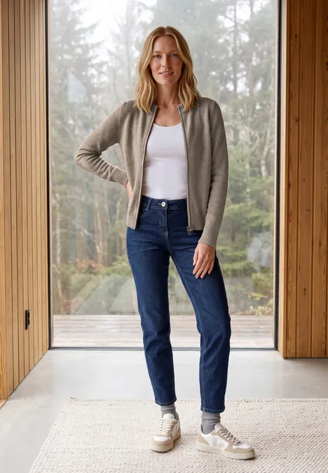 Eine Frau steht vor einem großen Fenster, trägt weiße Tanktop, graue Strickjacke, blaue Jeans und Sneaker.