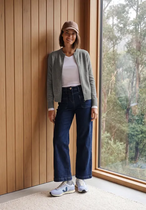 Frau in Baseballcap, grauem Cardigan, weißem Top und weiten Jeans steht neben Fenster und Holzpaneelen.